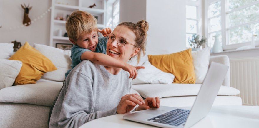 Geldanlage – Mutter mit kleinem Sohn sitzt in Wohnzimmer vor Laptop